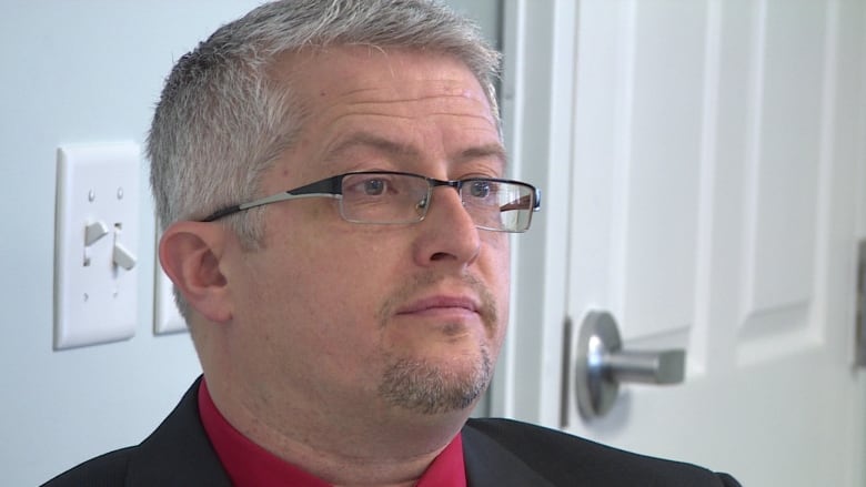 A close up of a man's face, who is wearing a black blazer and red tie.