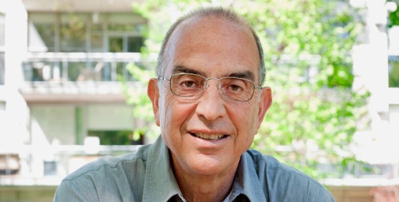 A man with glasses sits outside facing the camera. 