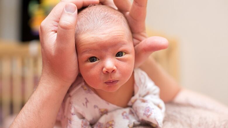 A young baby looks straight at the camera. Adult hands support its head.