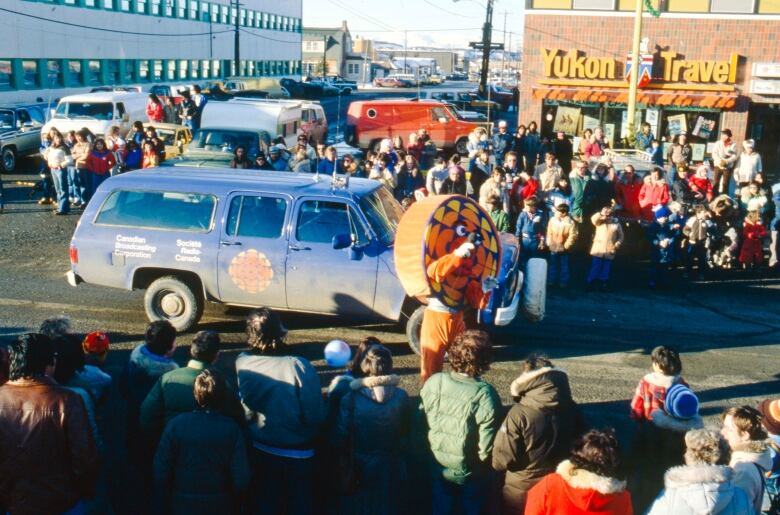 A CBC truck and a person in a CBC logo costume walk down a street in a parade as crowds look on from the sidewalk.