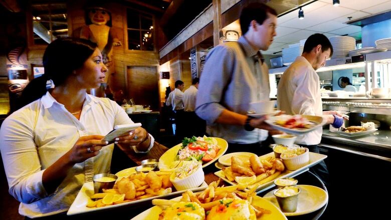 A woman with a large tray of full plates in front of her holds a small notepad. Another person holds a plate behind and to her left, while a third person, in the background on the right, appears to be placing something on a plate.