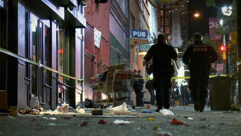 A store in Vancouver Downtown Eastside is a crime scene with yellow tapes sealing its entry. Piles of garbage on the ground and two police officers walking in the background.