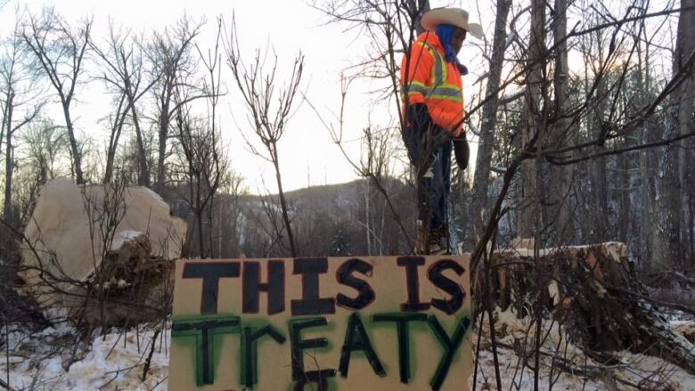 A man wearing a high-vis vest stands on a sign reading 'This is Treaty Land' on a snowy forest.