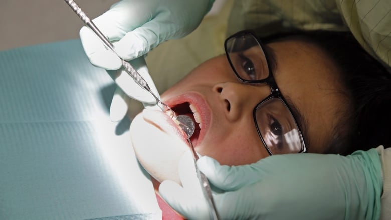 A young person wearing glasses is shown in a dentist's chair, with tools in their mouth.