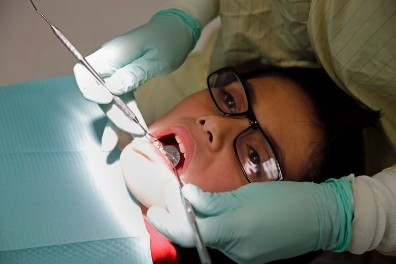 A young person wearing glasses is shown in a dentist's chair, with tools in their mouth.