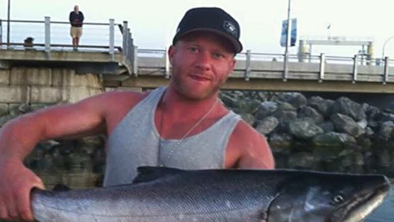 A white man with ruddy skin, wearing a black ballcap and a grey tank top, holds a large fish in both hands. 