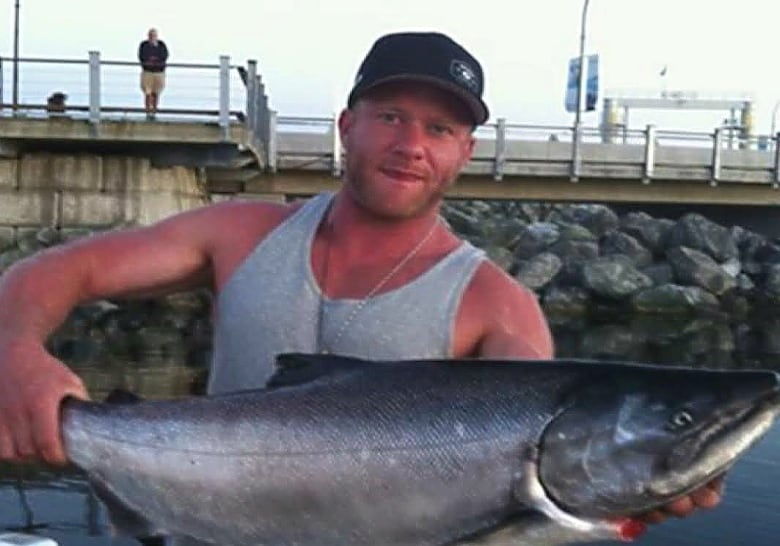 A white man with ruddy skin, wearing a black ballcap and a grey tank top, holds a large fish in both hands. 