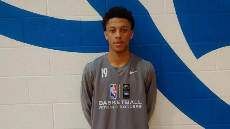 A young Black man wearing a grey jersey with the NBA logo that reads 