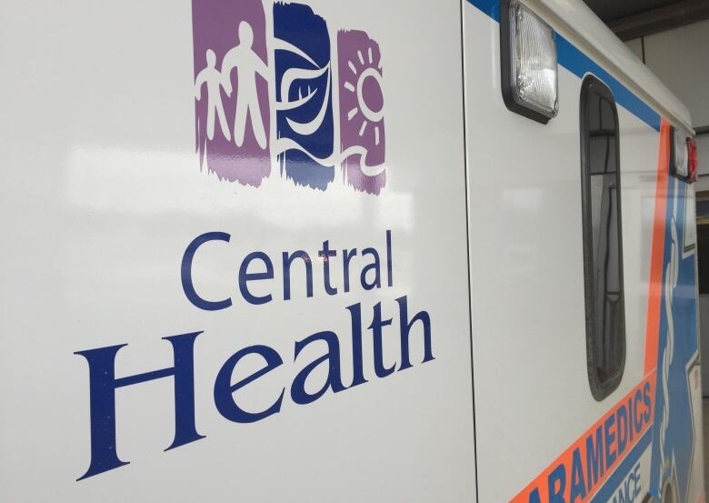 A closeup of the Central Health logo on the sign of a white ambulance. The logo shows a stylized rendering of two figures holding hands, a leaf and the sun above some waves. To the left of the logo the word 