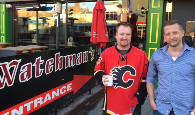 A man in a red Calgary Flames hockey jersey and another man in a blue button-up shirt stand beside the Watchman's Pub patio in Calgary.