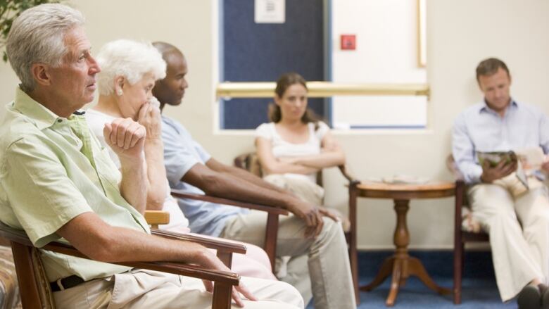 Five adults, including two seniors in the foreground, sit waiting in a doctor's office waiting room.