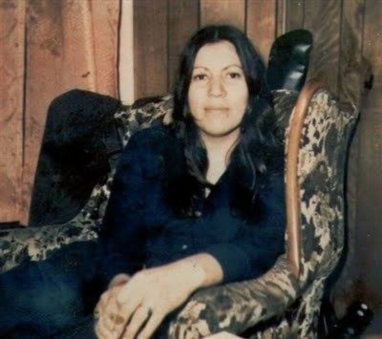 A woman with long black hair sits in an armchair looking into the camera.