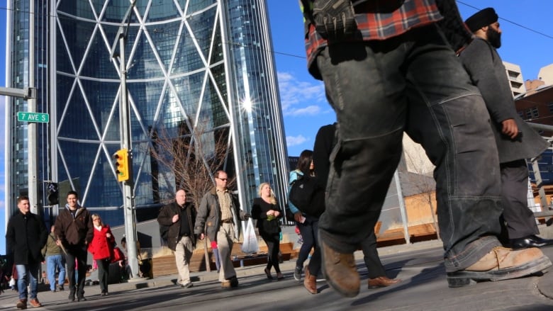 People wearing work boots and business attire walk across a busy downtown crosswalk. In the background is a large office tower. 