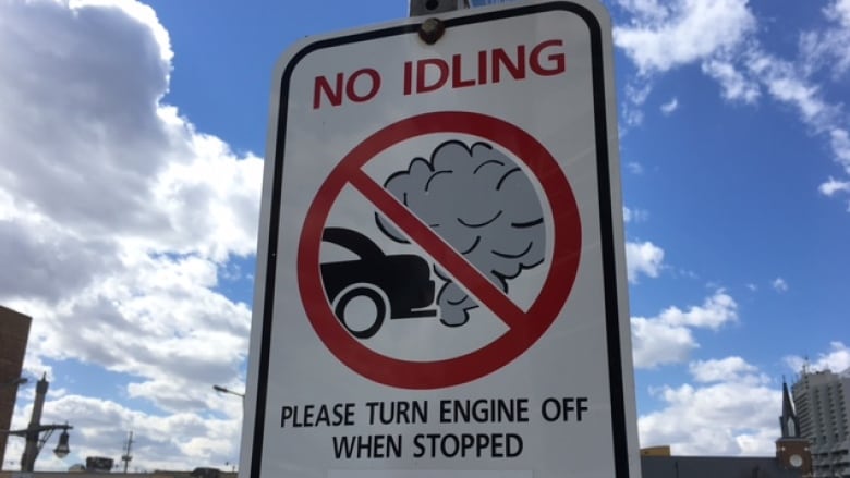 A white road sign with an image of a black car releasing emissions and a red circle with a line through it says 