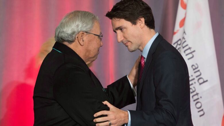 Two men shake hands in front of a flag that reads 'Truth and Reconciliation.'