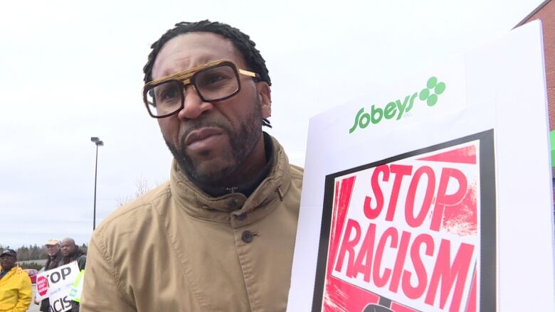 A Black man wearing glasses is seen holding a sign that reads 