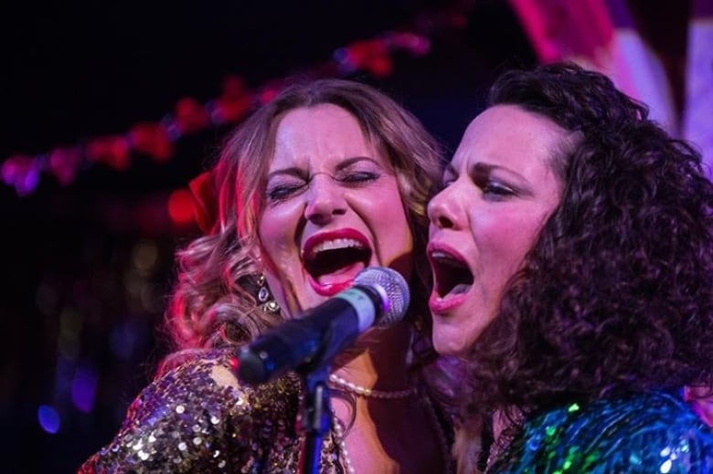 Two women on stage singing closely to one microphone. 