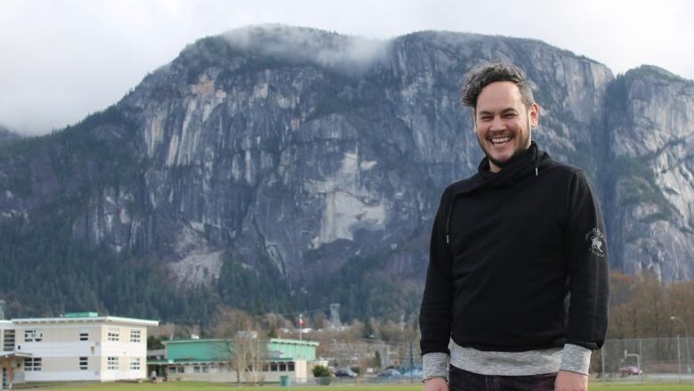 A man with a smile stands in front of a mountain and some buildings. 