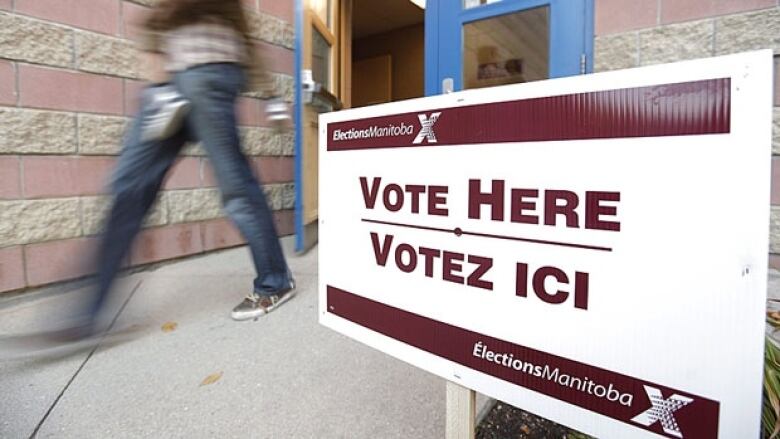 A person's legs are seen as they exit a door. Near them is a sign in the foreground that says Vote Here.