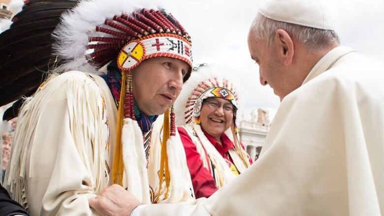 A man in a First Nations headdress shakes hands with a man in white papal robes.