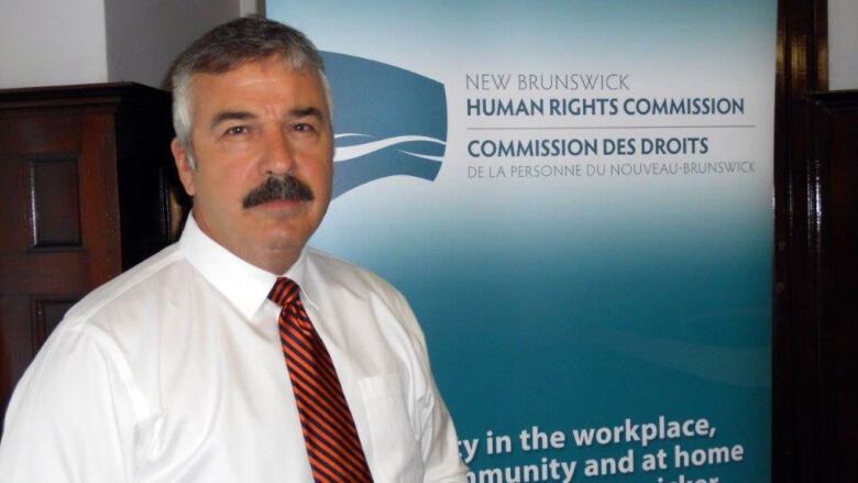 A man in a white shirt and a red and black-striped tie stands in front of a poster that says New Brunswick Human Rights Commission.