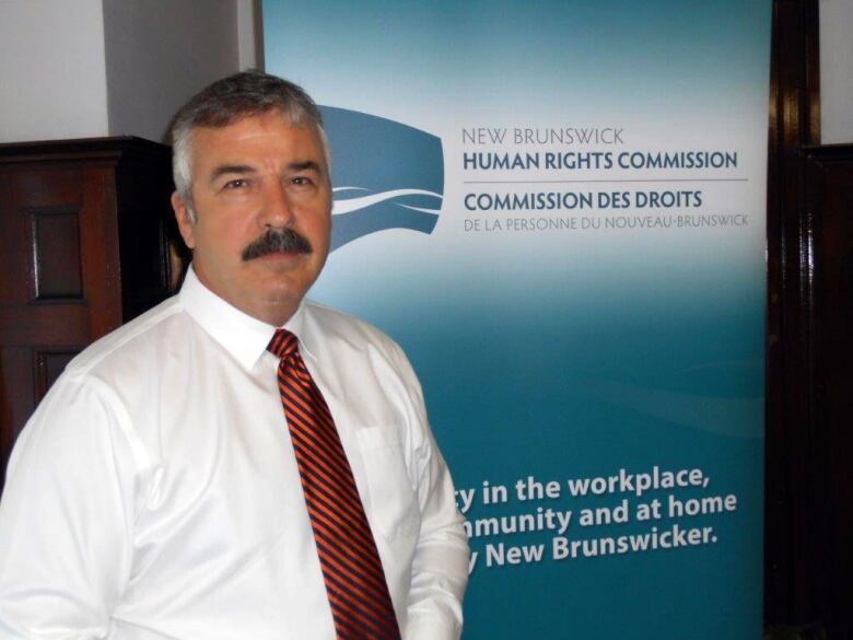 A man in a white shirt and a red and black-striped tie stands in front of a poster that says New Brunswick Human Rights Commission.