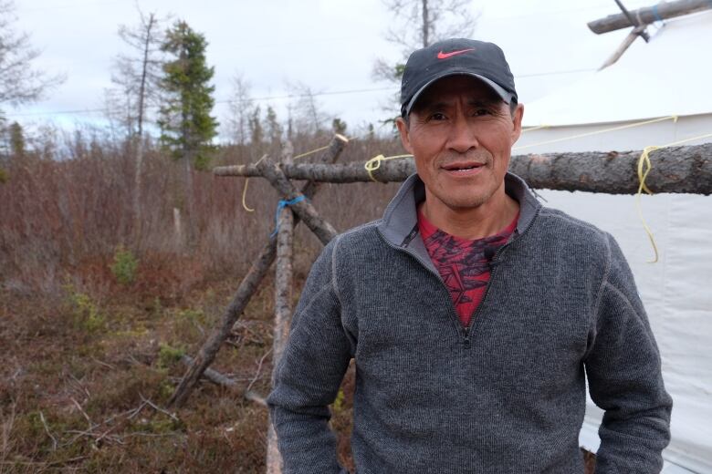 Man in sweater and hat stands in front of water and bush.