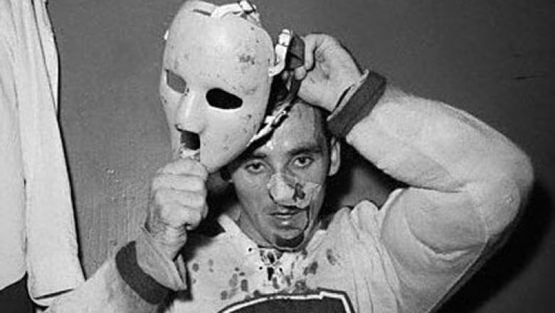 In this black-and-white photo, Jacques Plante puts on a white fiberglass mask. His nose is bandaged and his jersey has blood spots on it.