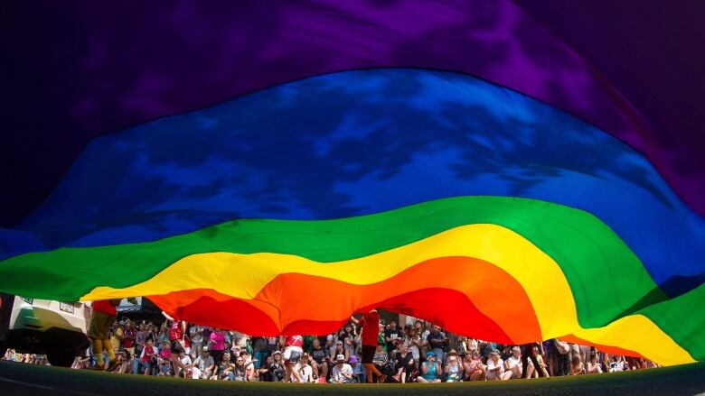 A series of people are visible beneath a large rainbow flag, which takes up the majority of the image.