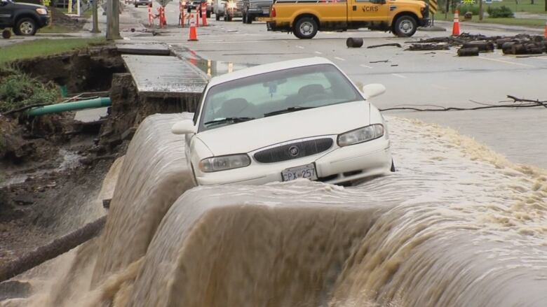 A car at an angle on the side of a washed-out road as flood water pushes it toward the edge.