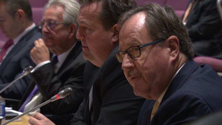 A row of men in suits sit before long microphones