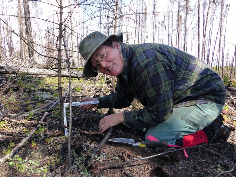 A women crouching in the soil uses an L-shaped silver tool dug next to some branches