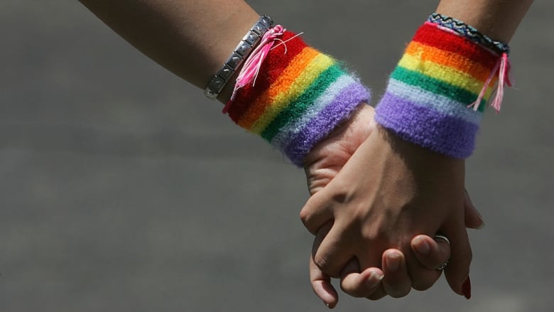 Two people hold hands in front of a gray background while wearing rainbow wristbands. 