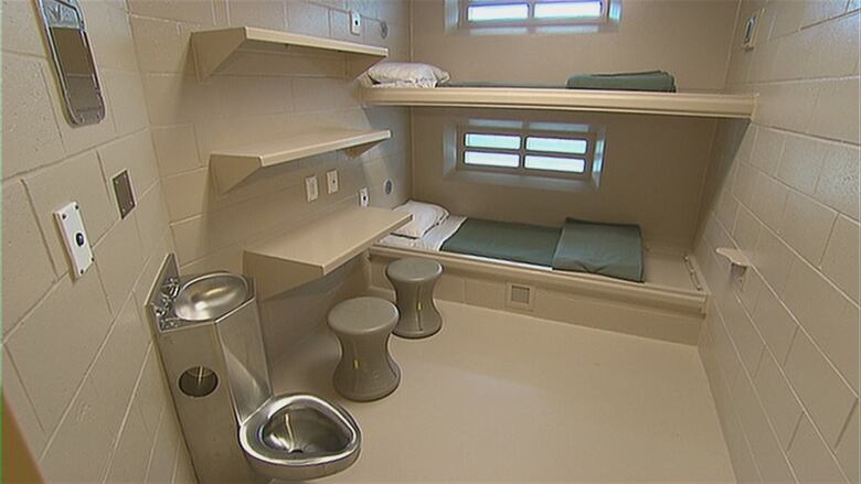 An empty jail cell is shown, including bunk beds, a sink and toilet, and two stools with a desk. The cell is located in the Southeast Regional Correctional Centre in Shediac, New Brunswick.