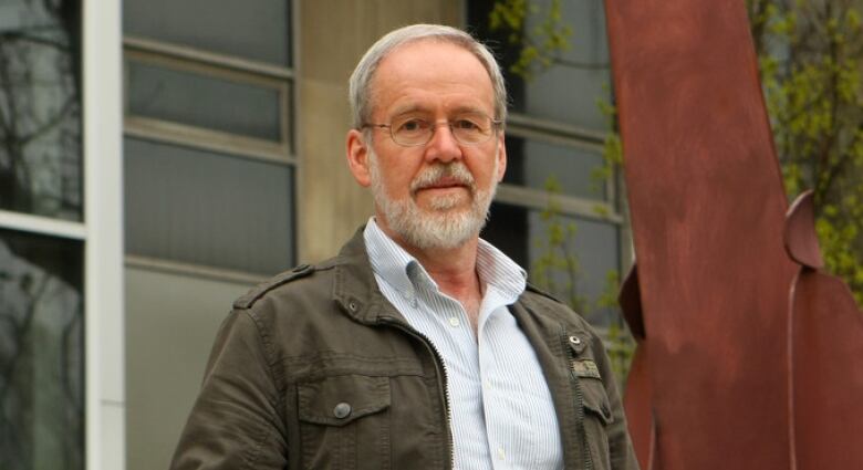 A man with a white beard and glasses stands outside a building.