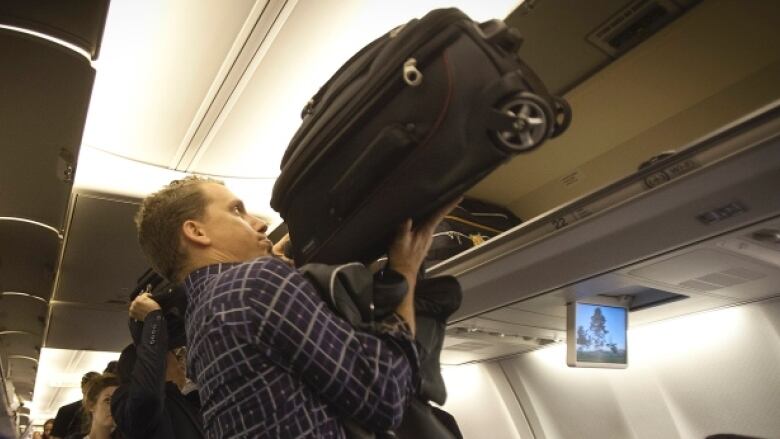 A man lifts a small black suitcase into the overhead bin in an airplane cabin.