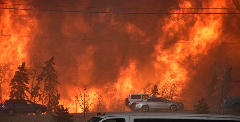 A large wild fire is blazing behind two trucks on the side of the road.