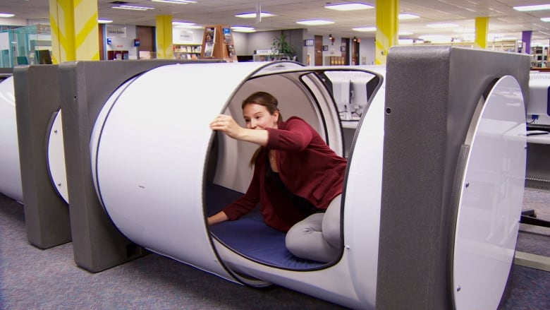 A woman climbs inside a white cylindrical pod located inside a college library.