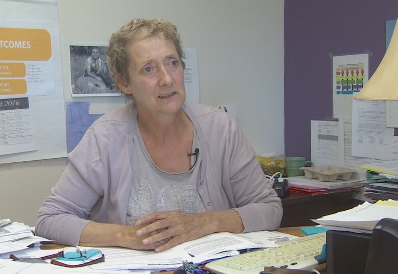 A woman is pictured speaking with sitting behind a desk covered with papers.