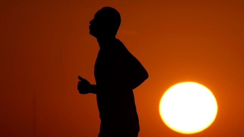 A silhouette of a person jogging under the gaze of an orange sky and bright sun.