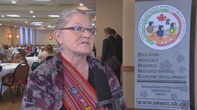 A woman is photographed standing next to a banner for the Saskatchewan Aboriginal Women's Circle Corporation.