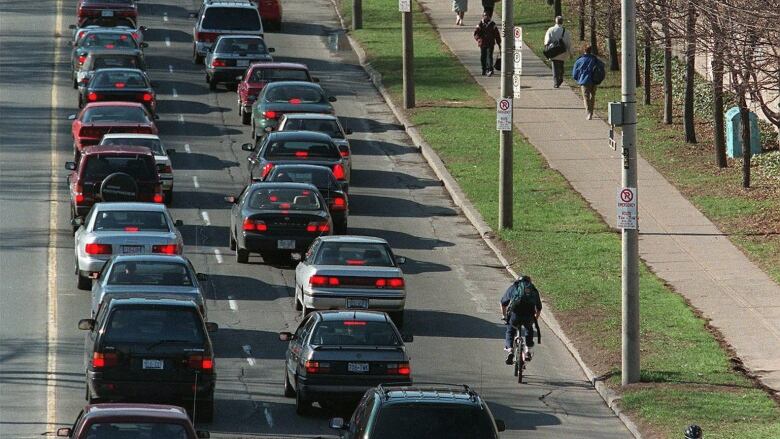 Cars are lined up bumper to bumper on a long, straight road. Some bikers and pedestrians are passing them on the right.