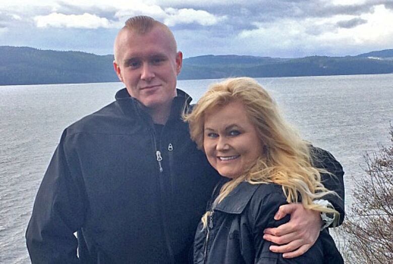 A young man in a black jacket has one arm over his mother, a woman in a black coat with long, blonde hair. They smile at a camera, behind them a body of water, mountains in the horizon.