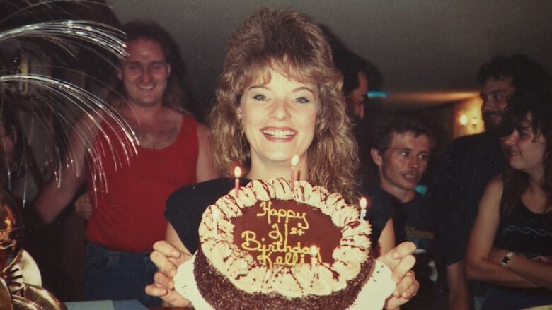 People stand behind a young woman as she smiles and holds a cake with the words 'Happy 21st Birthday Kelly' on it.