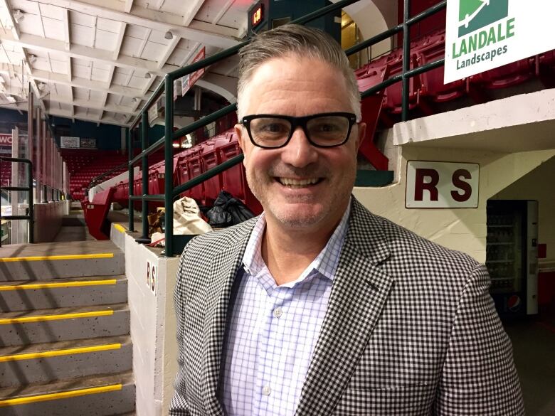 A man wearing glasses and a suit jacket smiles at the camera in a hockey arena