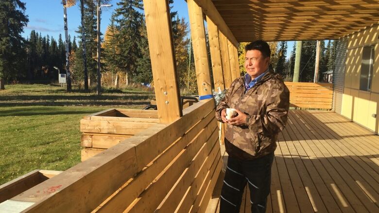 A man stands squint in the sun on a wooden deck. He is warming a camo jacket.