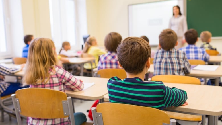 The back of students' heads can be seen as they listen to a teacher at the front of a classroom.