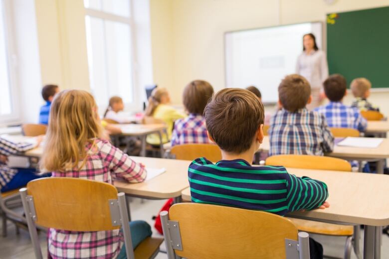 The back of students' heads can be seen as they listen to a teacher at the front of a classroom.