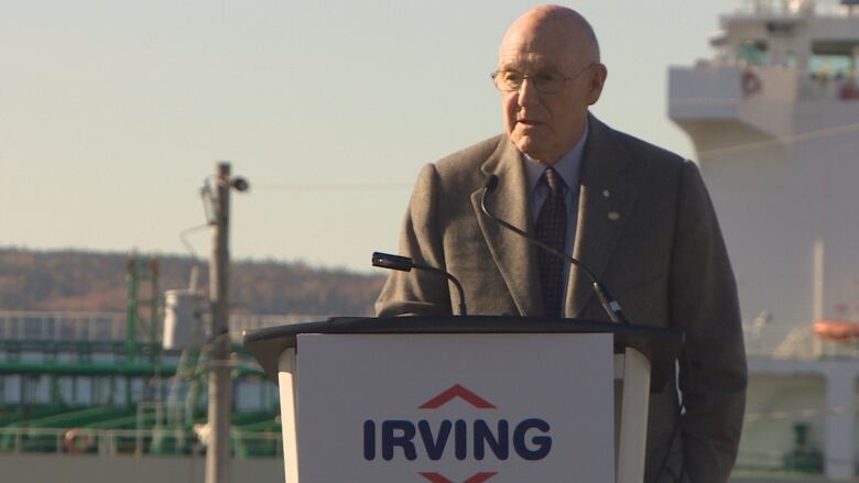 An older man wearing a business suit speaks outdoors at a lectern that features the Irving corporate logo on its front. 