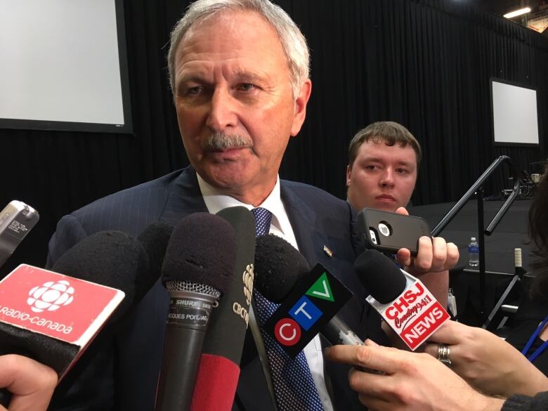 A man with short grey hair and a dark grey moustache talks to a variety of media microphones held in front of him.